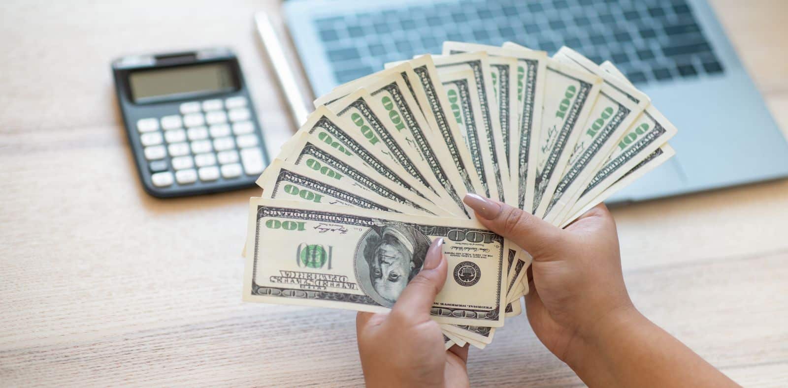 A hand offers a folded stack of $100 bills to a smiling young woman in a pink cardigan against a bright pink background
